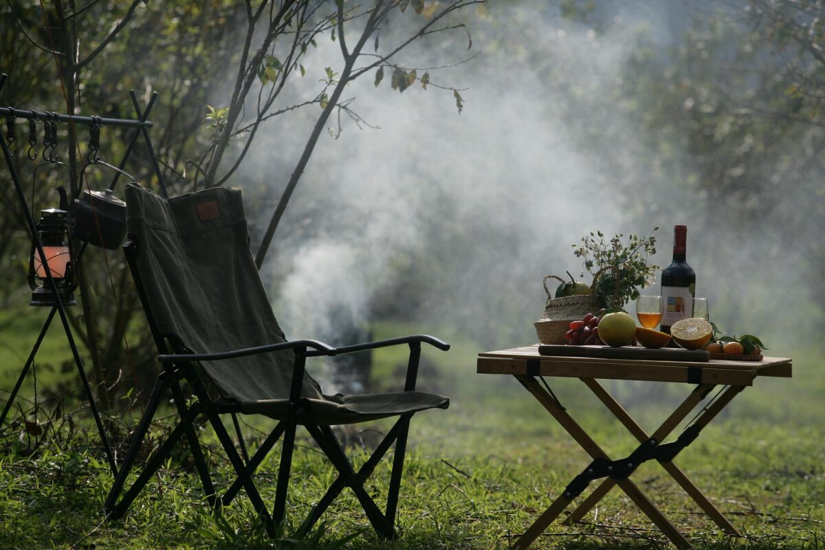 Camping chair for one safari guest with smoke from a fire in the background. On a small wooden table, wine, flowers, and fruit.