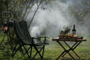 Camping chair for one safari guest with smoke from a fire in the background. On a small wooden table, wine, flowers, and fruit.